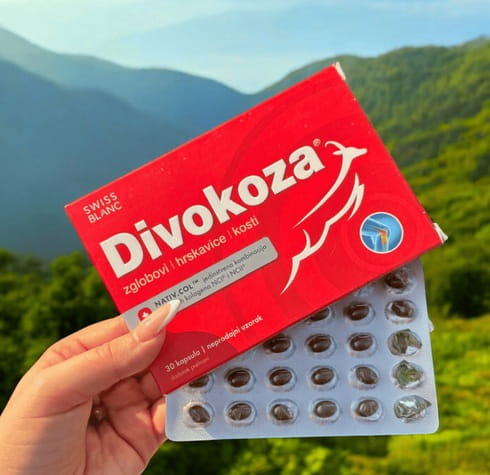 Ženska ruka drži kutiju Swiss Divokoza i blister s kapsulama ispred zelene planinske krajolike.